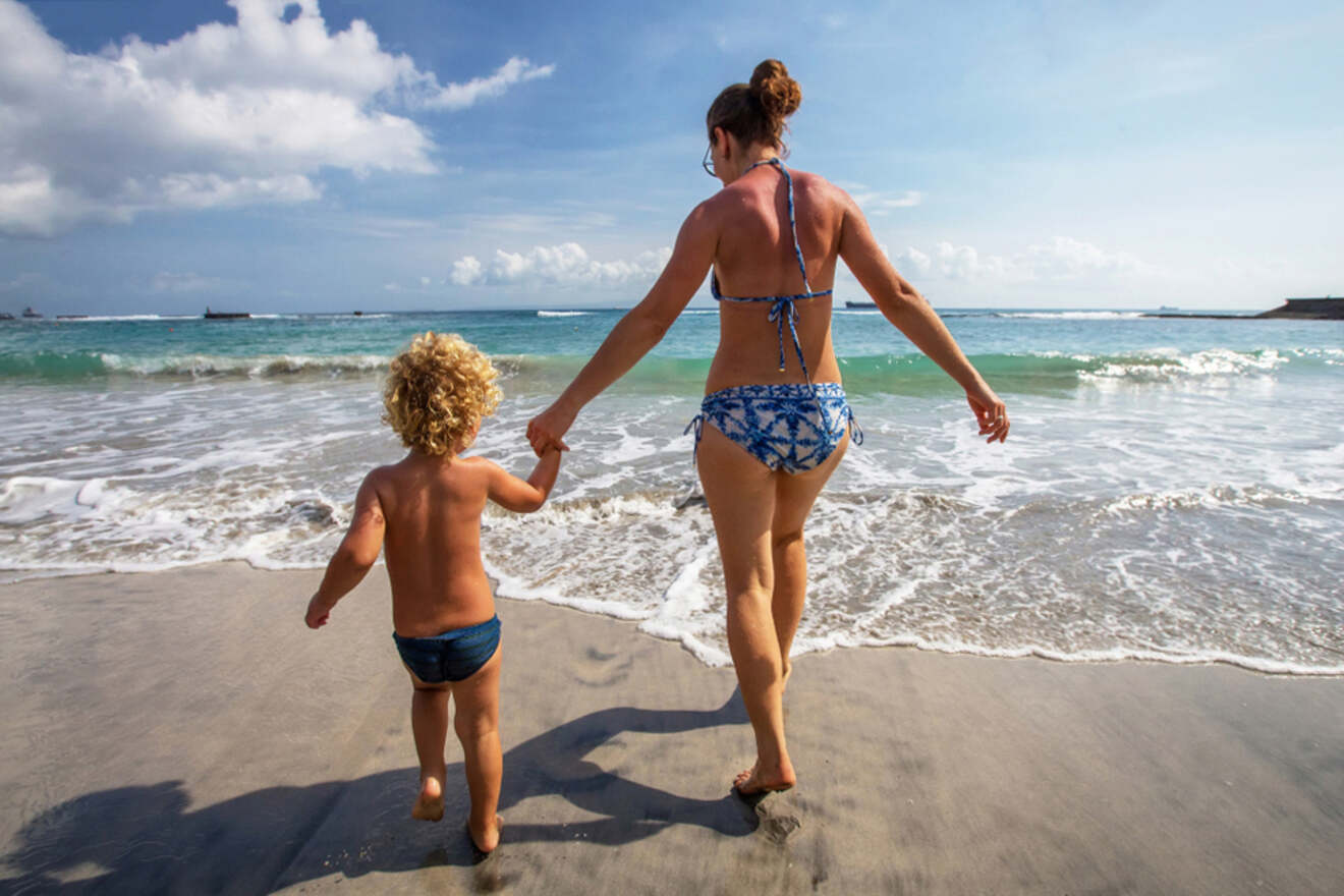 An adult in a blue bikini walks hand in hand with a child in swim trunks along the shore of a sandy beach with gentle waves under a clear sky.