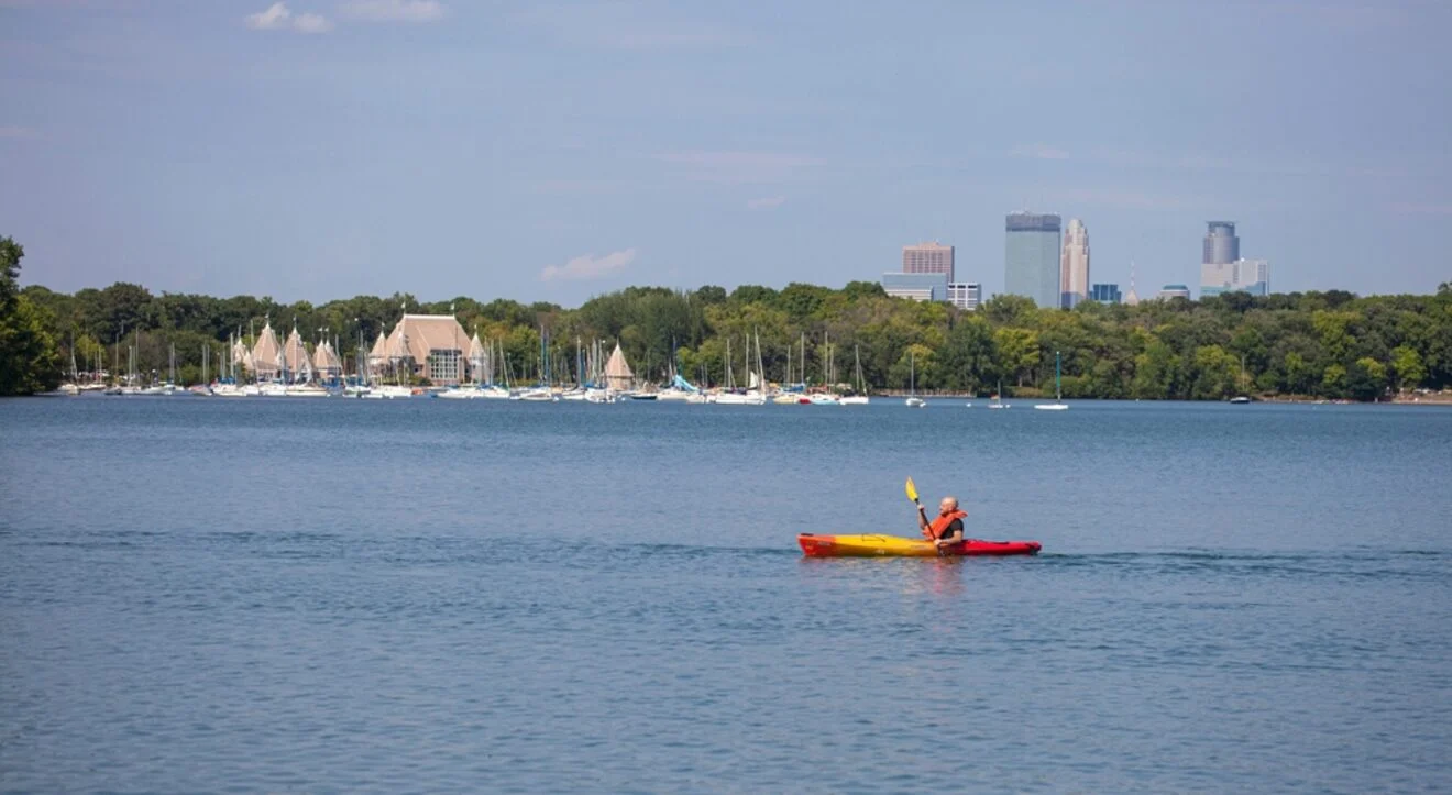 A person kayaks on a calm lake with sailboats docked in the background and a city skyline visible in the distance under a clear blue sky.