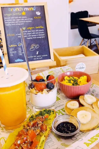 A brunch spread featuring avocado toast, a yogurt parfait with berries, orange juice, scrambled eggs with chives, and mini pancakes topped with banana slices and syrup.