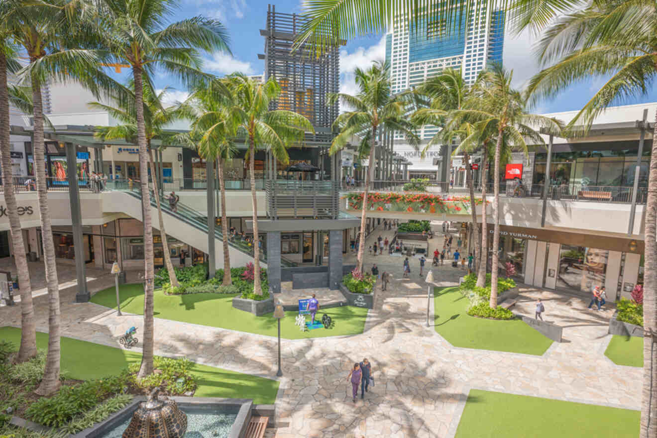 Outdoor shopping mall with palm trees, upper and lower level stores, and people walking. Sunny day with modern architecture and a fountain in the foreground.