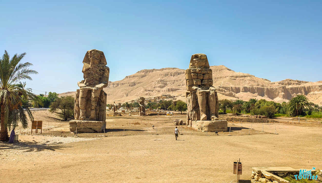 Two massive stone statues, the Colossi of Memnon, stand against a desert landscape with distant mountains, surrounded by sparse vegetation. A person walks nearby for scale.