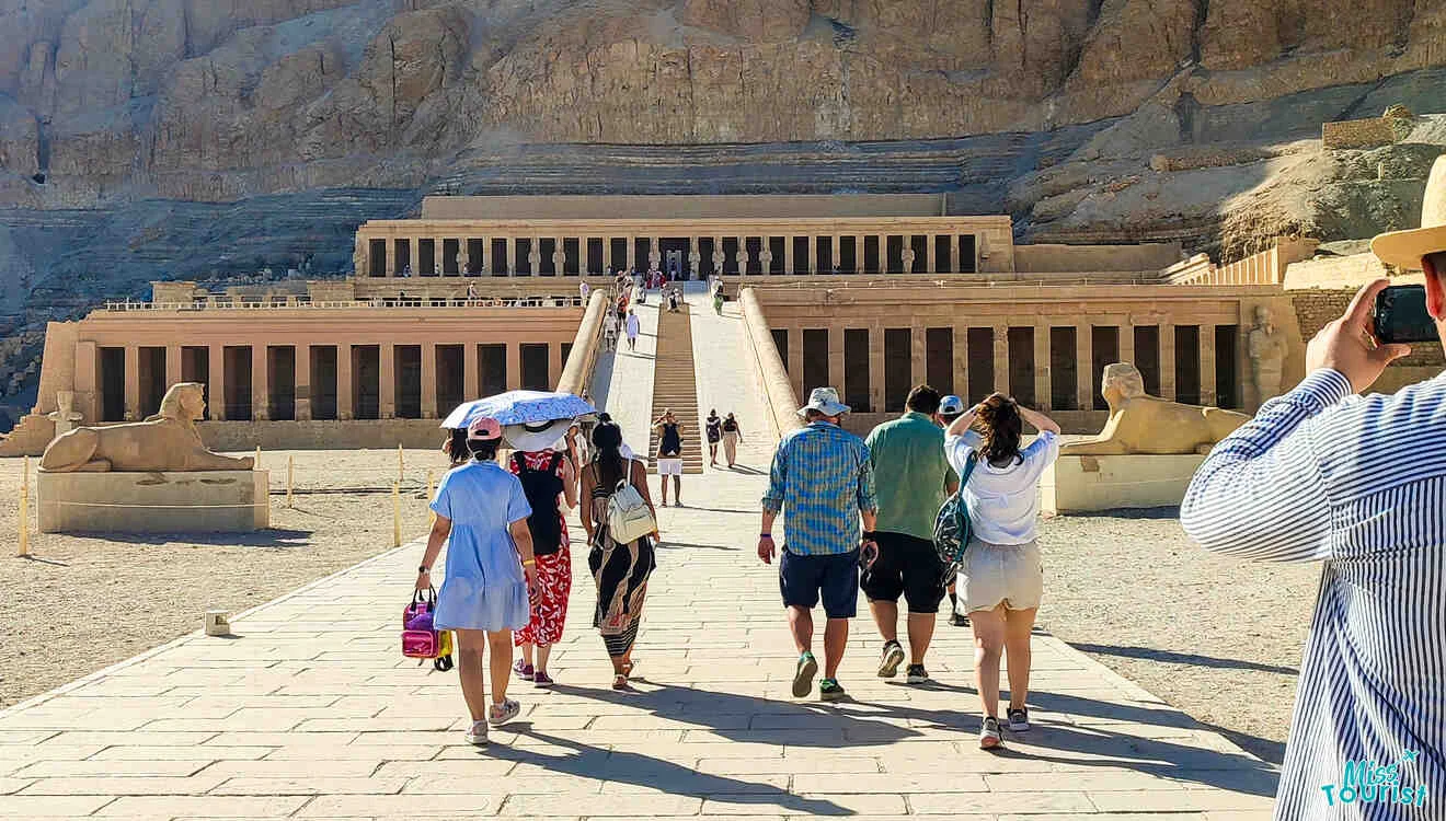 Tourists walk toward the ancient Egyptian temple of Hatshepsut, with stone statues lining the staircase.
