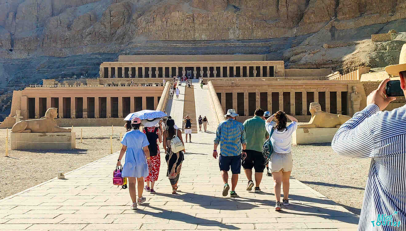 Tourists walk toward the ancient Egyptian temple of Hatshepsut, with stone statues lining the staircase.