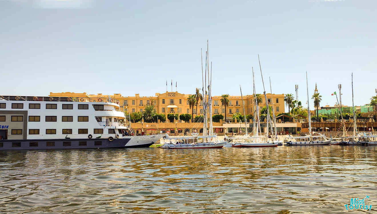 A large riverboat and several sailboats are moored alongside a sunlit river with a beige building and palm trees in the background.