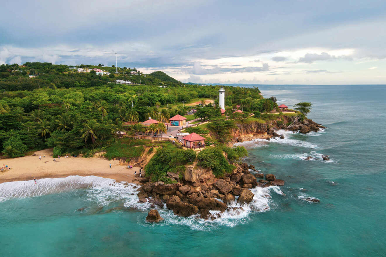 Aerial view of a coastal area with a lighthouse, surrounded by greenery and rocky cliffs. A small beach with people is visible along the shoreline, with the ocean in the background.