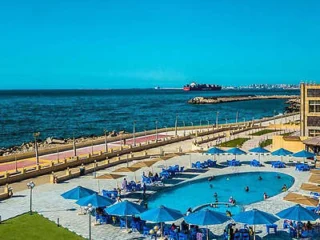 Coastal scene with a circular pool surrounded by blue umbrellas and chairs. People are swimming. A large ship is visible on the ocean, with a distant shoreline under a clear blue sky.