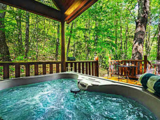 Outdoor hot tub on a wooden deck with a forest view. A rolled towel is placed on the tub edge, and a small round table with chairs is visible in the background.