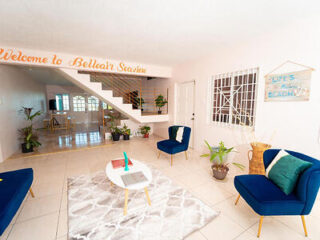 Bright living room with blue chairs, a round white table, and plants. "Welcome to Belleair Seaview" sign on wall. Stairs lead to upper floor. Wall decor reads "Life's a Beach.