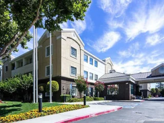 A three-story beige building with a pitched roof and landscaped entrance, surrounded by trees and flowers under a blue sky with wispy clouds.