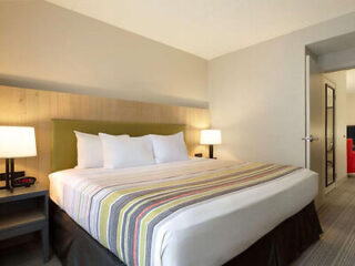 A hotel room with a neatly made bed, striped bedspread, two bedside tables with lamps, and an open door to an adjacent room. Walls are light-colored and the decor is minimalistic.