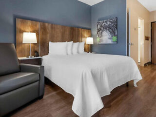 Hotel room with a white bed, two bedside lamps, a dark armchair, a landscape photo on the wall, and wood flooring.