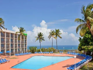 Outdoor hotel pool area with palm trees, surrounded by blue lounge chairs, near a multi-story building. Ocean view in the background under a clear blue sky.