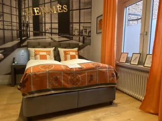 A bedroom with an Hermès theme, featuring a bed with orange and white bedding, matching pillows, a bedside table, framed pictures, and large windows with orange curtains.