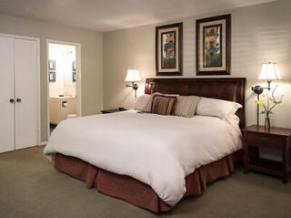 Bedroom with a large bed, brown headboard, and two nightstands with lamps. Decor includes framed art above the bed and a vase with a flower. A bathroom is visible through an open door.