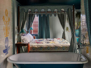 Four-poster bed with patterned quilt next to a freestanding bathtub in a vintage-style room with tiled walls and curtains.