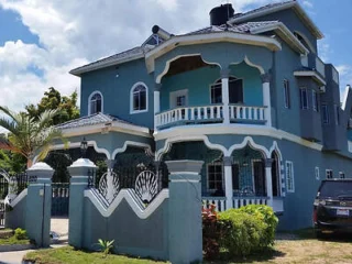 Two-story blue house with white trim, featuring a balcony and decorative railing. A black car is parked in front, and a palm tree is in the yard. Cloudy sky in the background.