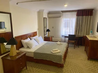 Hotel room with a double bed, two wooden side tables with lamps, a desk, chair, TV, and a window with curtains. Towels are folded on the bed.