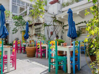 Colorful outdoor cafe with blue umbrellas, bright chairs, and tables surrounded by plants and a white building background.