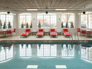 Indoor pool with red lounge chairs, tables, and large windows. Ceiling features wood paneling, and the pool is surrounded by tiled flooring.