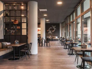 Modern restaurant interior with wooden tables and black chairs, large windows on the right, and decorative wall art in the background.