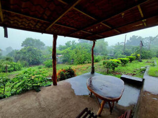 Covered porch with a round wooden table and bench overlooking a lush, green garden on a foggy day.