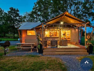 A lit-up glamping tent with a wooden deck, picnic table, and grill in the foreground, surrounded by trees at dusk.