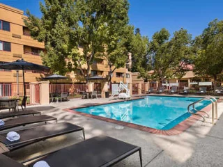 Outdoor swimming pool surrounded by lounge chairs and shaded tables, adjacent to a multistory building and trees.