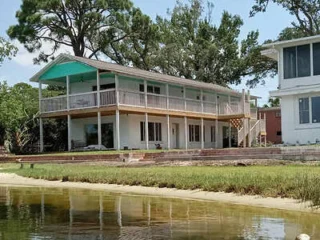 A two-story house with a large wraparound balcony overlooks a calm water body. It is surrounded by trees and has a grassy area in the foreground.
