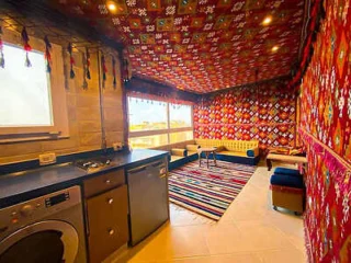 Room with colorful patterned walls and ceiling, a striped rug, a small kitchen area with appliances, and a seating area with couches and a table.