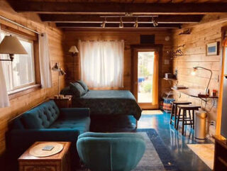 Cozy wooden cabin interior with a bed, teal sofa, armchair, desk, and various light fixtures. Large windows and a door provide natural light.