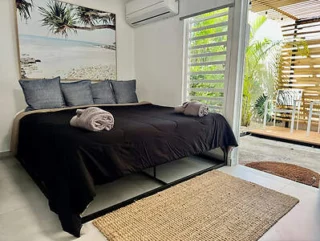 A bedroom with a large bed, black bedding, and two rolled towels. There's a beach painting on the wall, an air conditioner above, and an outdoor area with wooden slats visible through a door.