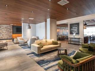 Modern lounge with beige couches, green armchairs, and a blue rug. Stone accent wall and wall art featuring text. Recessed lighting and wood-paneled ceiling. Dining area in the background.