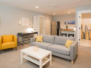 A modern living room with a gray sofa, yellow chair, and a white coffee table. A small desk is in the corner. The kitchen area with a fridge and a bar counter is visible in the background.