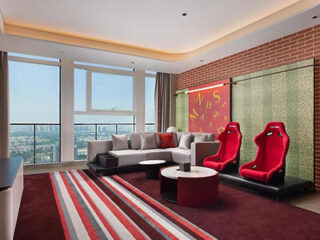 A modern living room with a city view, featuring a large sofa, two red racing-style chairs, a patterned rug, and a decorative wall with red and yellow accents.