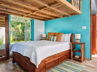 A cozy bedroom with a wooden bed, white bedding, and turquoise accent wall. The ceiling features exposed wooden beams. Two bedside tables with lamps are visible, and a window opens to a garden view.