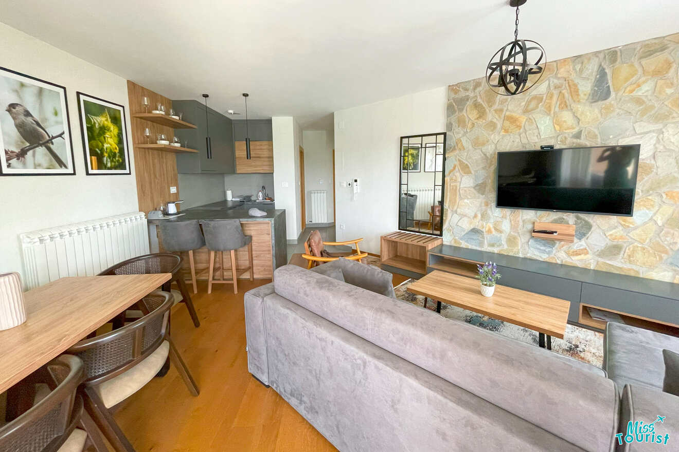 Experience the perfect blend of style and function in this modern living room and kitchen, featuring gray couches, a wooden dining table, a wall-mounted TV, and captivating artwork—ideal for unwinding after planning your next Serbia itinerary.