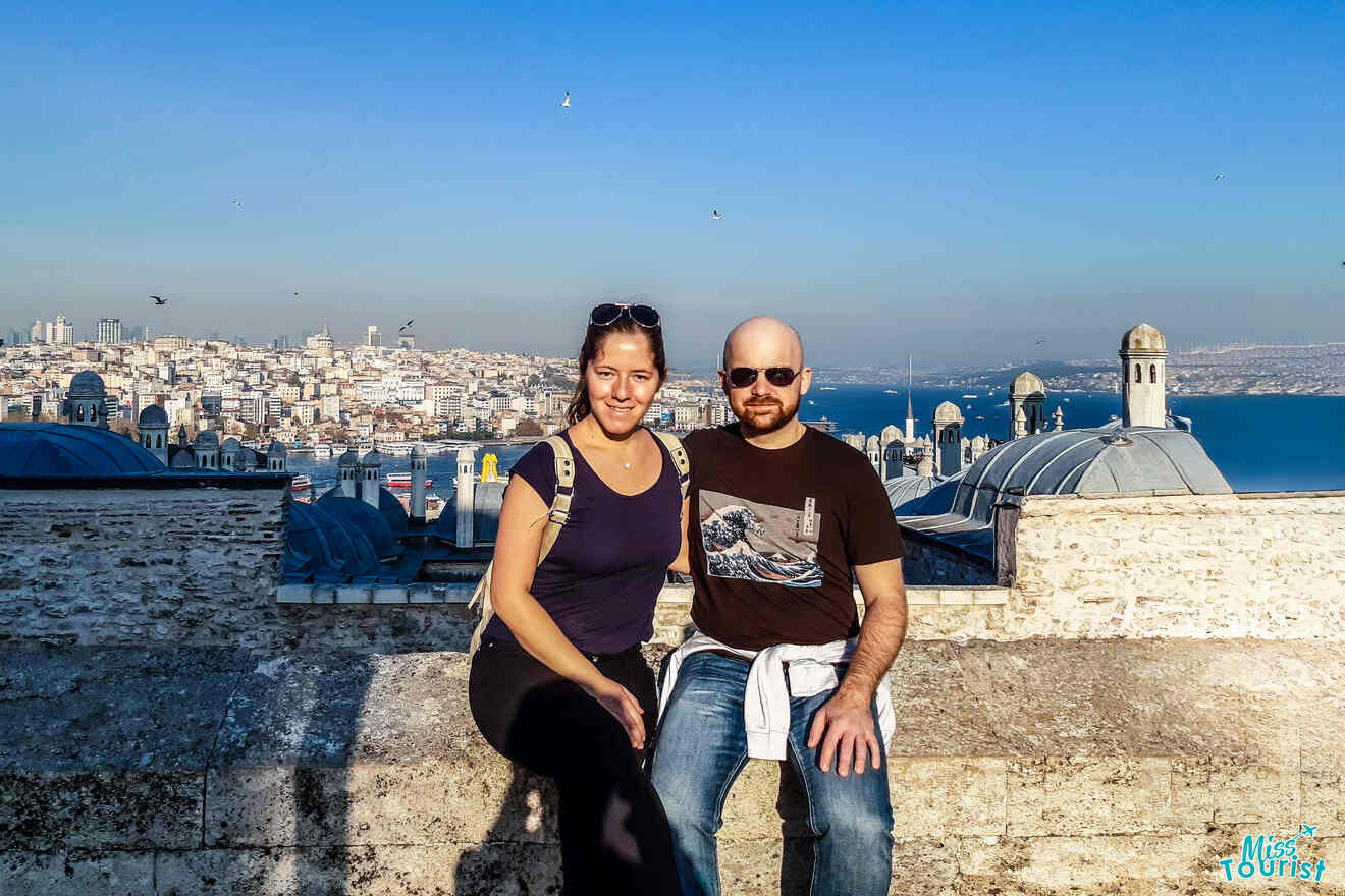 The writer of the post with her husband sitting on a stone wall with a cityscape and blue sky in the background.