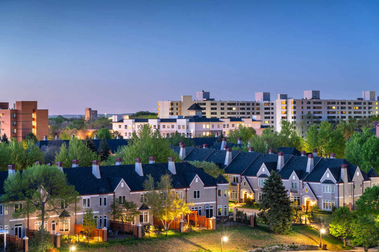 Residential area with two-story houses and trees in the foreground, with multi-story apartment buildings in the background under a clear evening sky.