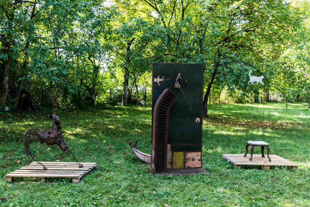 Outdoor art installation featuring a metal horse sculpture, a central abstract door-like structure, and a stool sculpture, all placed on wooden platforms in a lush green park setting.