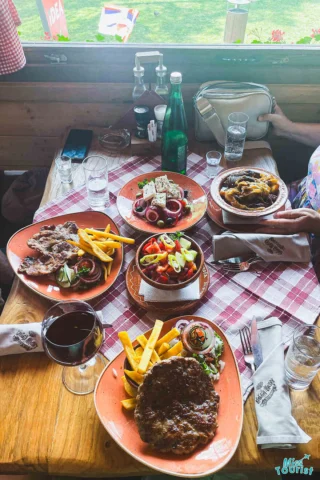 A welcoming table set with plates of grilled meat, vegetables, fries, and salad invites you to explore the flavors of a Serbia itinerary. There are drinks, including red wine and a green bottle, all atop a checkered tablecloth.