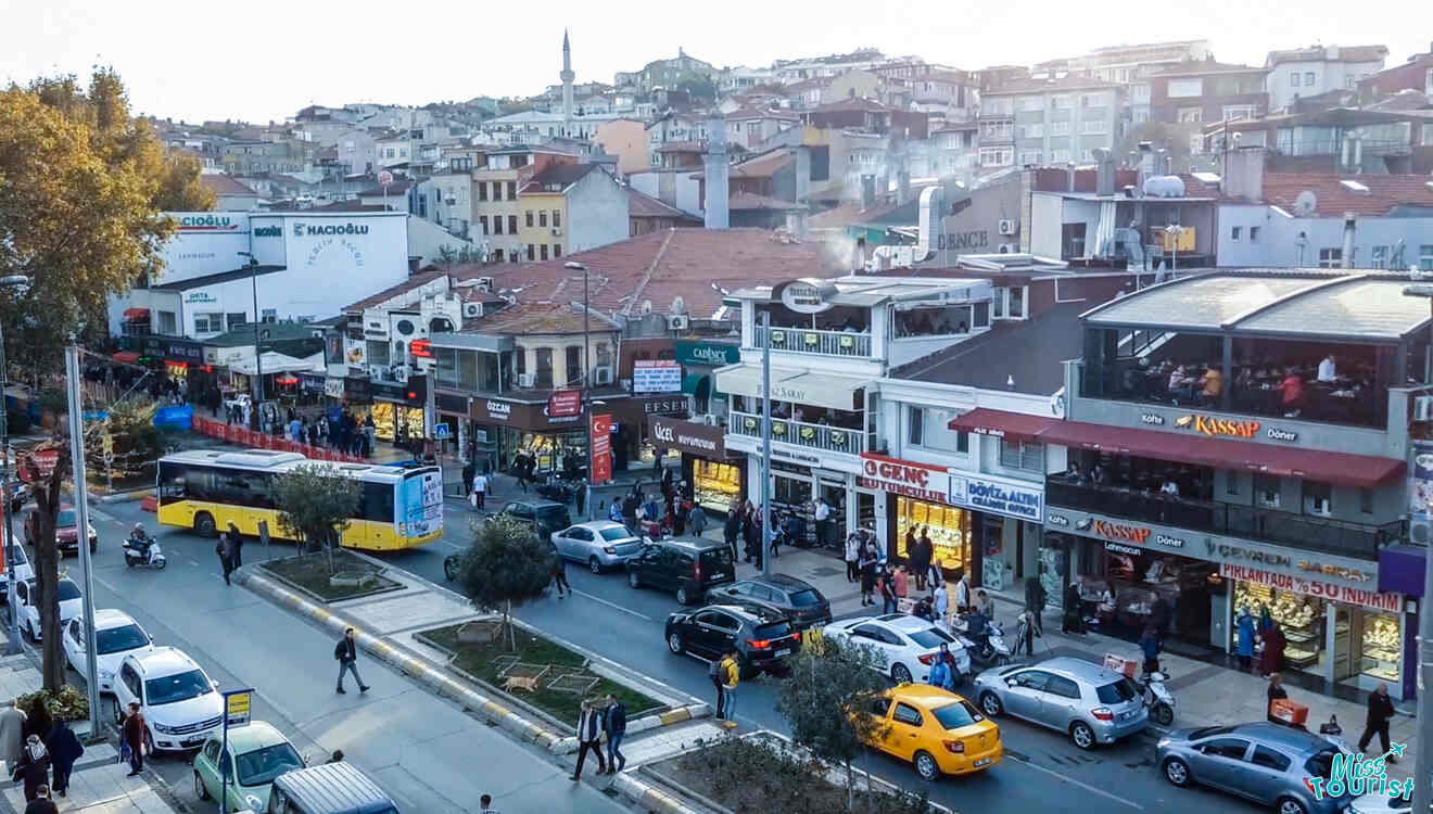 A busy street scene in a city with shops, a bus, and cars. People walk along the sidewalks. Buildings with various signs are visible in the background.