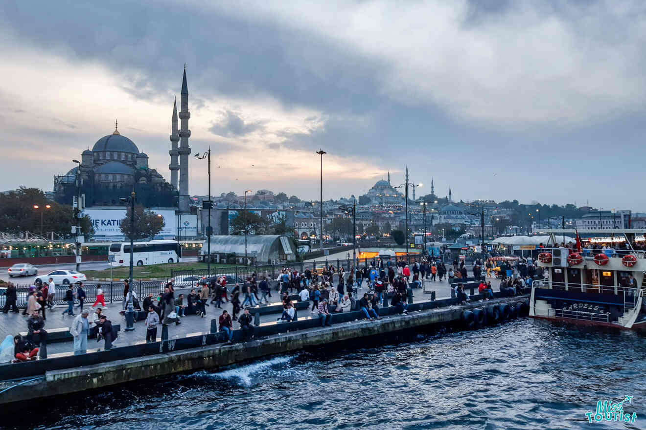 A bustling waterfront scene in Istanbul with people on a pier, a boat docked, and mosques with minarets in the background under a cloudy sky.