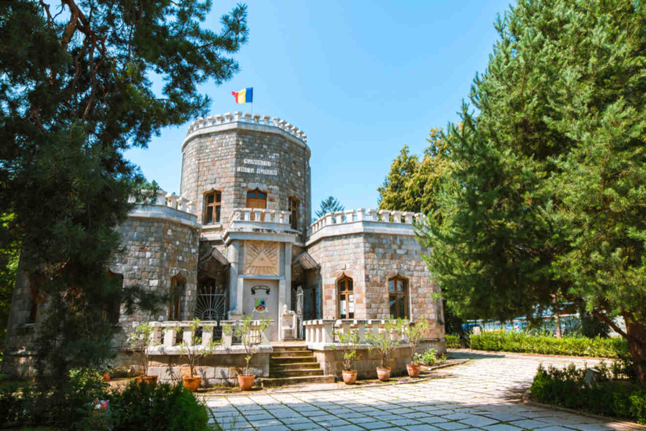 A stone building resembling a small castle with a round tower, surrounded by trees and a paved area. A flag is displayed on top.