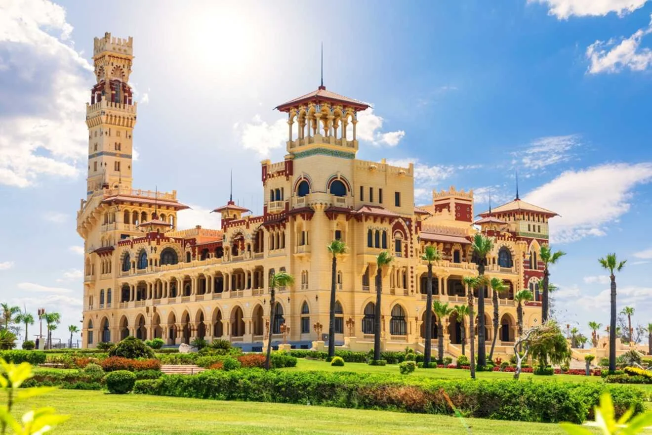 A grand palace, Al Montazah Palace, with ornate architectural details and a large tower, set in lush gardens under a blue sky with scattered clouds.