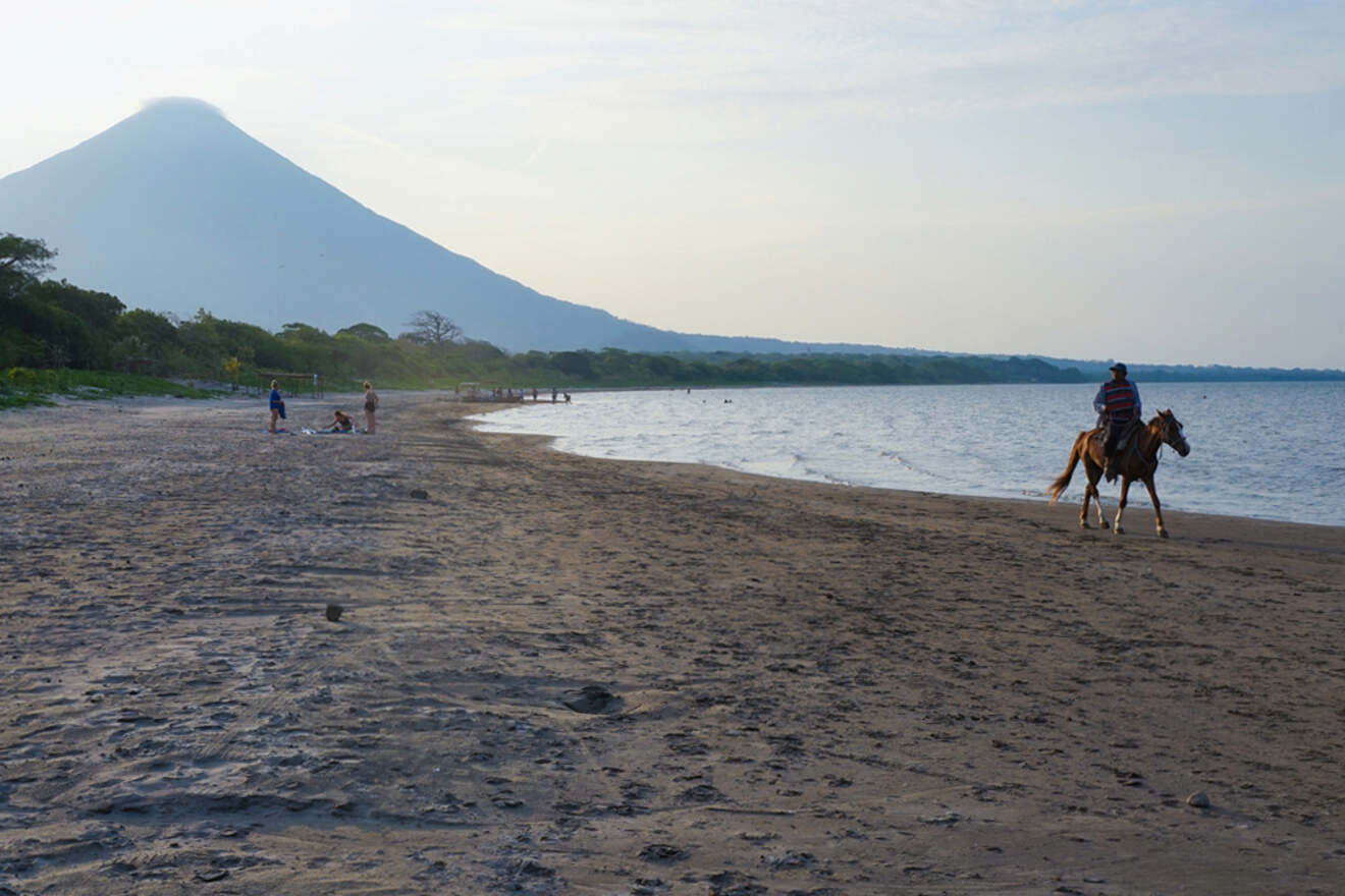 A person rides a horse along a beach with a volcano in the background. A few people are walking near the water's edge.