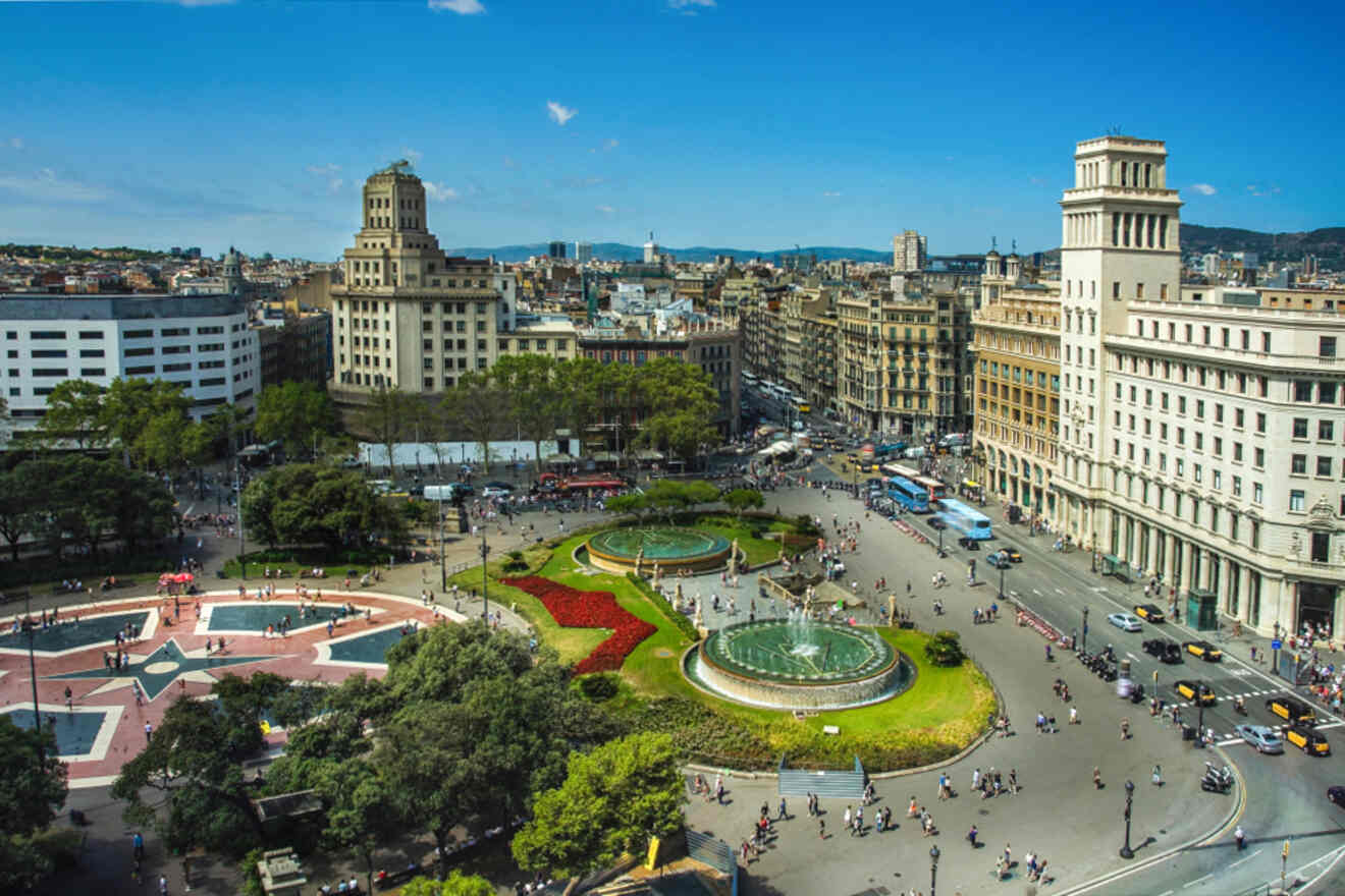 Aerial view of a bustling city square with two fountains, surrounded by historical buildings and trees, under a clear blue sky.