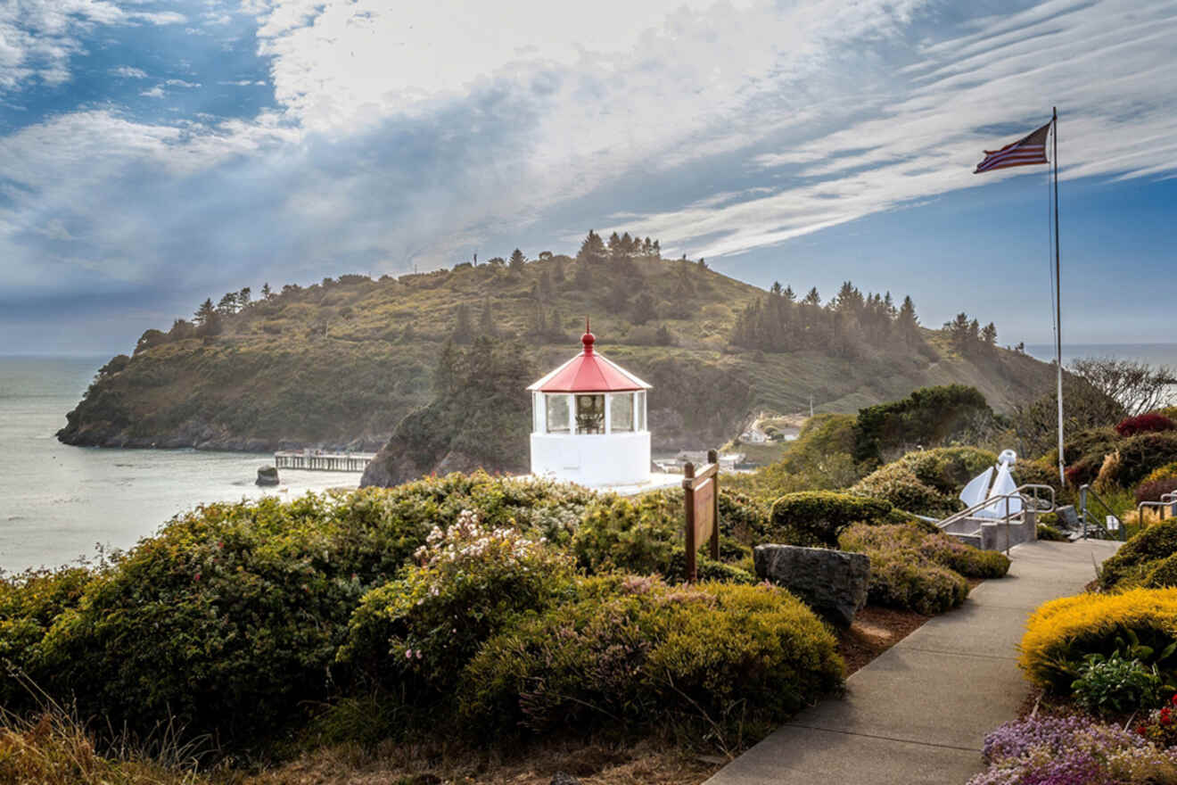 Coastal landscape with a white lighthouse, an American flag, and a pathway surrounded by greenery. The ocean and a forested hill are in the background under a partly cloudy sky.