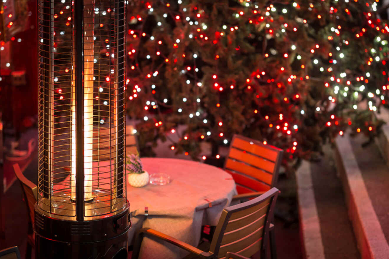 A patio heater is next to a table with chairs, illuminated by red and white Christmas lights on a nearby tree.