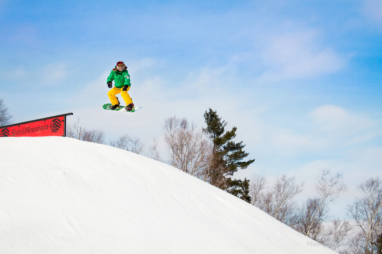 Person snowboarding off a jump with a clear blue sky and trees in the background.