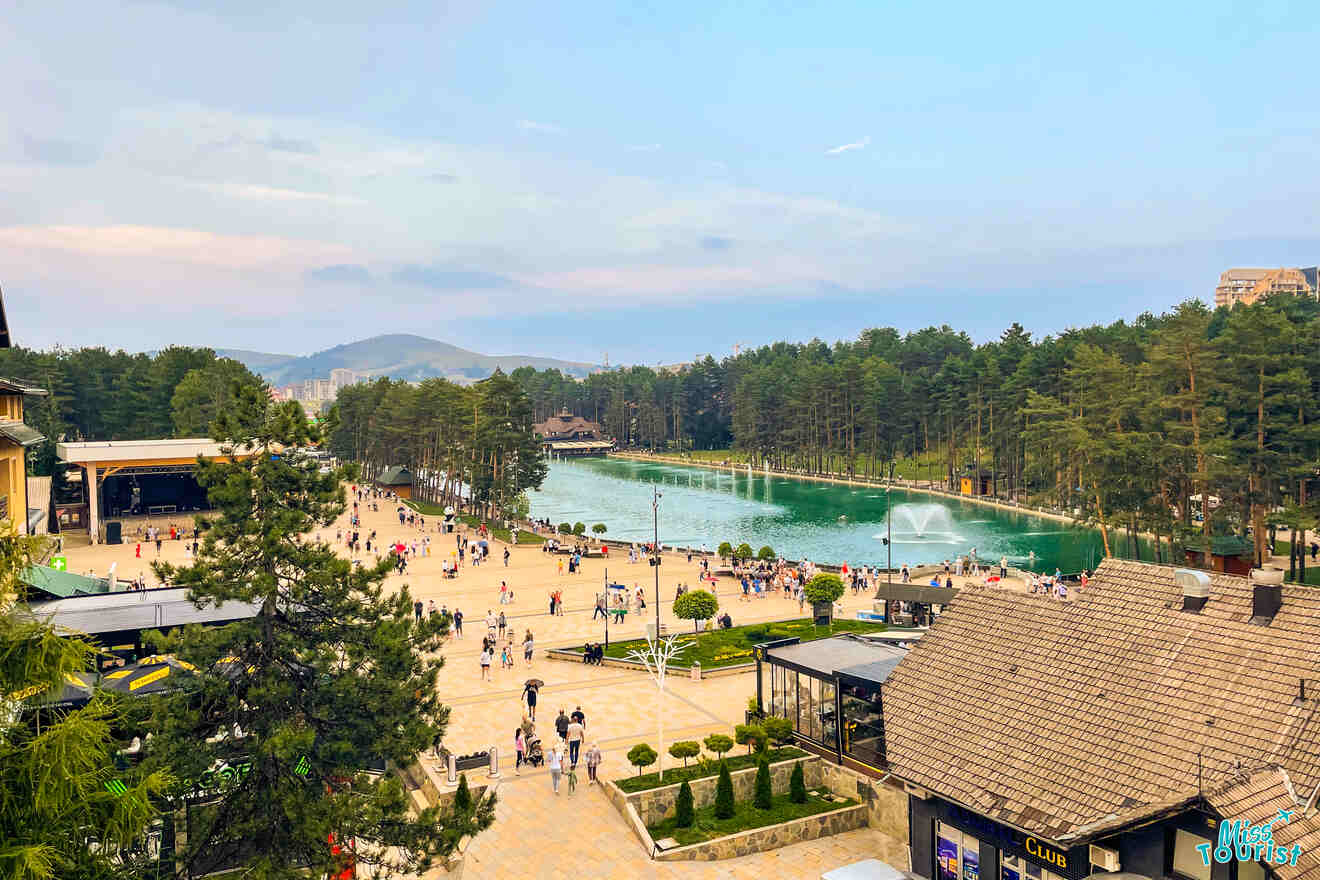 Aerial view of a scenic park in Serbia, perfect for your itinerary, with a lake surrounded by trees and people strolling along paved paths. A fountain graces the lake, framed by rolling hills in the background.